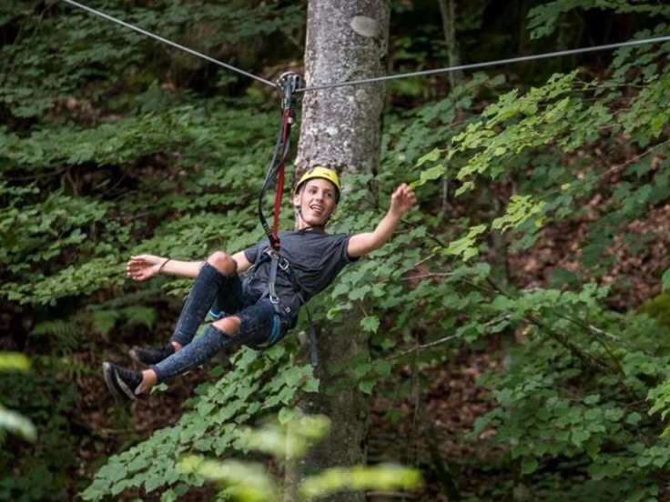 Une personne adepte de l'accrobranche, heureuse, accrochée à une tyrolienne, dans la forêt, à proximité du camping 3 étoiles avec piscine Le Petit Pyrénéen en Ariège, dans les Pyrénées