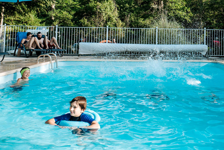Des adultes sur des transats discutent au bord de la piscine et des enfants nagent dans la piscine du camping 3 étoiles avec piscine Le Petit Pyrénéen en Ariège dans les Pyrénées